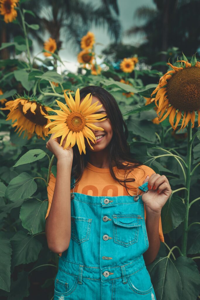 Joyful woman in denim holding sunflower among vibrant blooms in summer garden.